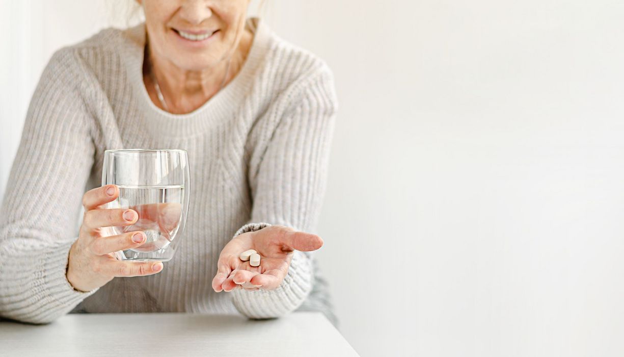 Ältere Frau, hält ein Glas Wasser und zwei Tabletten in der Hand.