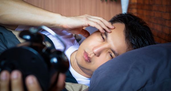 Ein schlafloser, junger Mann liegt nachts im Bett. Er schaut auf eine Uhr.