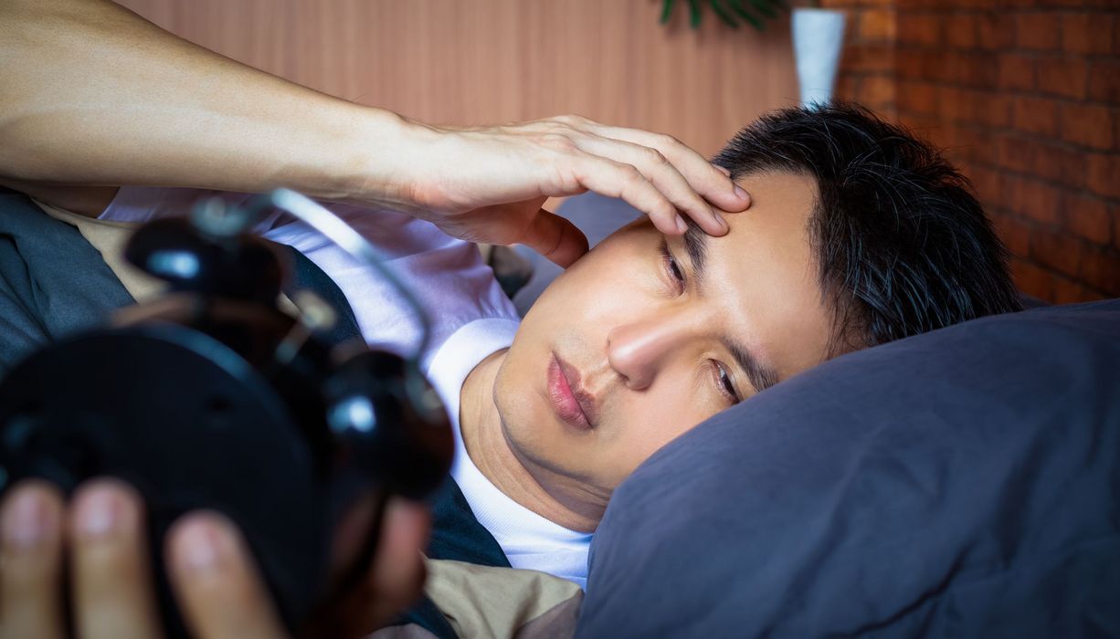 Ein schlafloser, junger Mann liegt nachts im Bett. Er schaut auf eine Uhr.