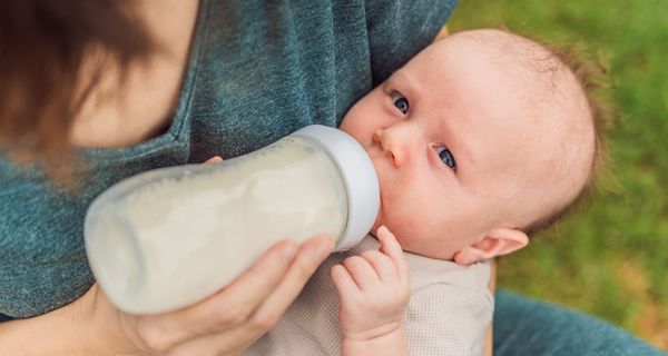 Baby trinkt auf dem Schoß einer Frau aus einer Flasche. Es blickt zweifelnd in die Kamera.