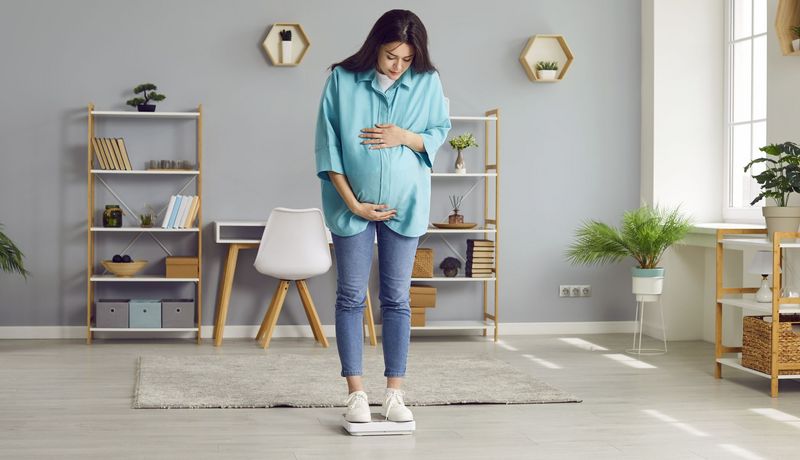 Schwangere Frau steht auf der Waage und blickt skeptisch auf das Ergebnis.