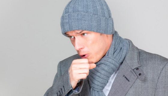 Mann mit grauer Mütze und grauer Jacke hustet und hält sich die Hand vor den Mund
