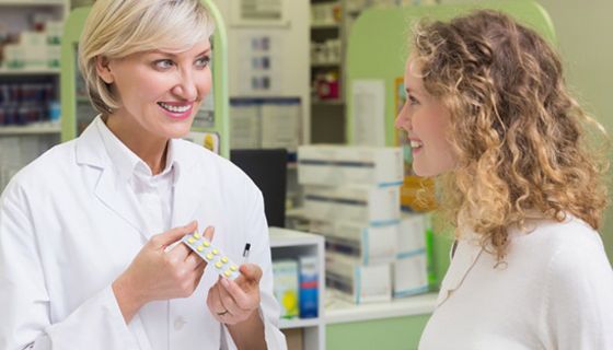 Beratungsgespräch: Apothekerin (in den 30ern, blond) mit Blister in der Hand berät Kundin in den 20ern (schulterlange, leicht gelockte, mittelblonde Haare). Beide lächeln