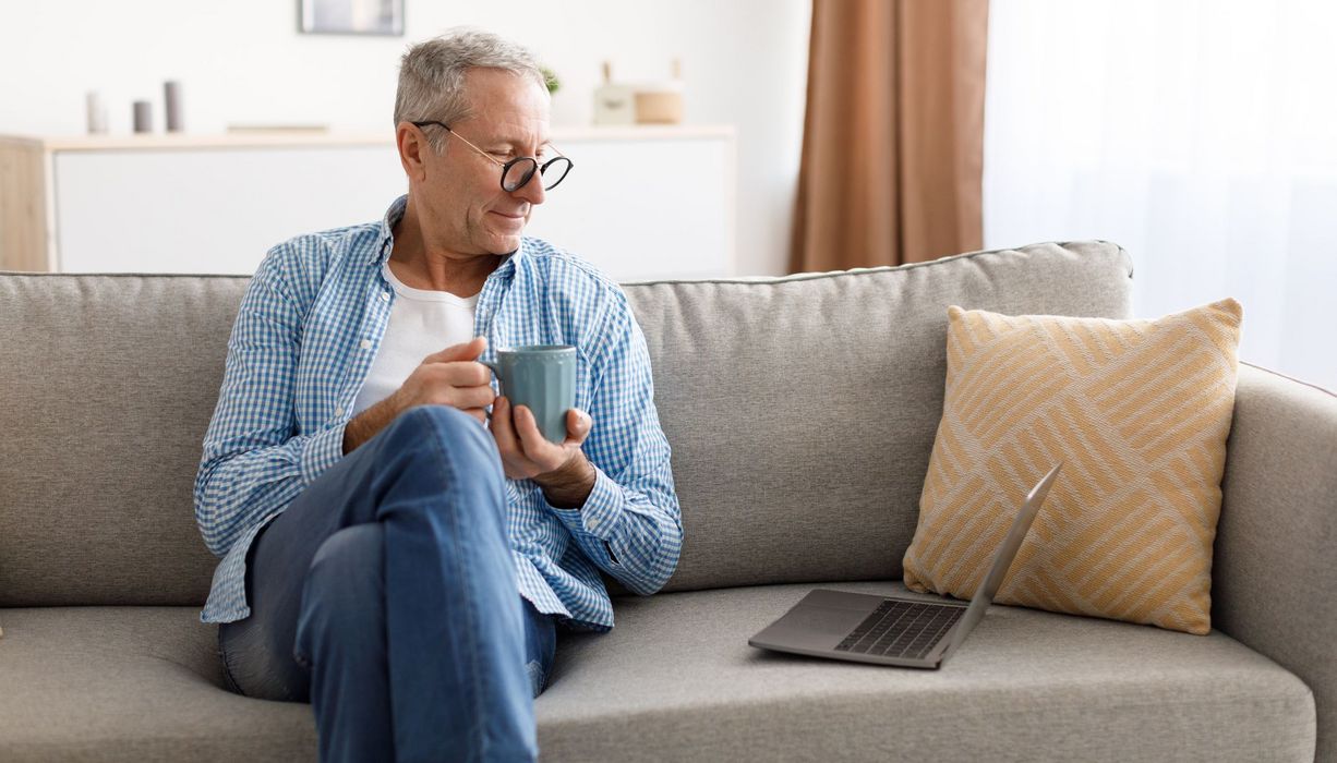 Älterer Mann, sitzt mit einem Kaffee auf der Couch, neben sich ein Laptop.