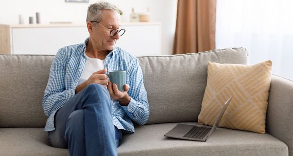 Älterer Mann, sitzt mit einem Kaffee auf der Couch, neben sich ein Laptop.