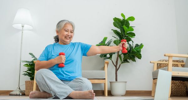 Eine ältere Frau sitzt in einem Wohnzimmer auf einer Sportmatte. Sie hat Hanteln in der Hand und schaut auf einen Laptop, der vor ihr steht.