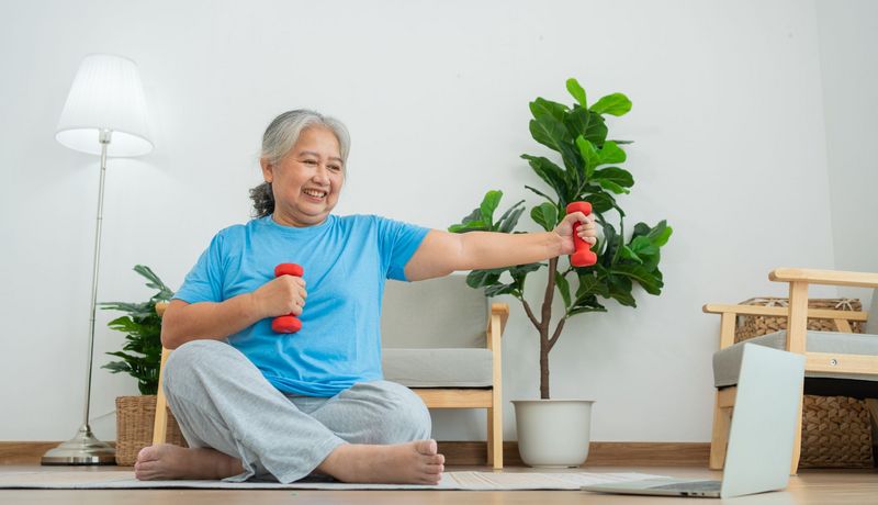 Eine ältere Frau sitzt in einem Wohnzimmer auf einer Sportmatte. Sie hat Hanteln in der Hand und schaut auf einen Laptop, der vor ihr steht.