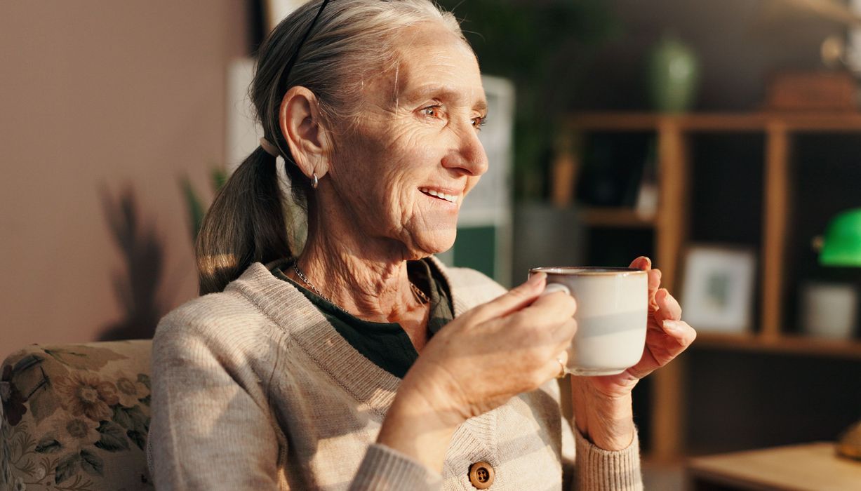 Eine Frau über 65 hält lächelnd eine Tasse in beiden Händen.