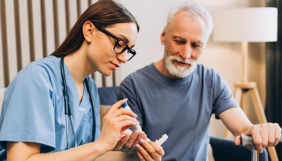 Eine junge Krankenschwester misst den Blutzucker eines älteren Herrn der neben ihr sitzt. 