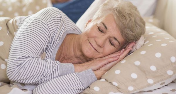 Eine ältere Dame schläft erholsam in einem Bett. Im Hintergrund ist ihr Ehemann zu sehen.