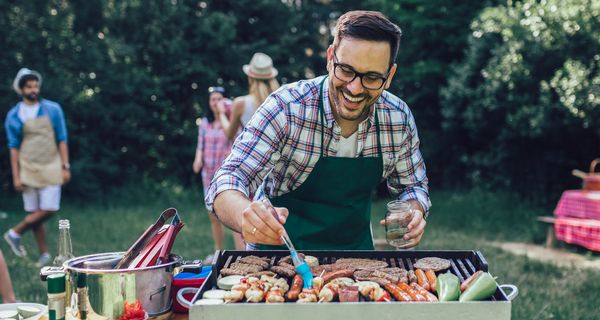 Mann bestreicht freudestrahlend einen Gemüsespies, der gemeinsam mit Zuchinischeiben, Paprika, Würstchen und Burgerpatties auf einem Grill liegt. Im Hintergrund sind unscharf ein paar gutgelaunte Personen zu sehen.