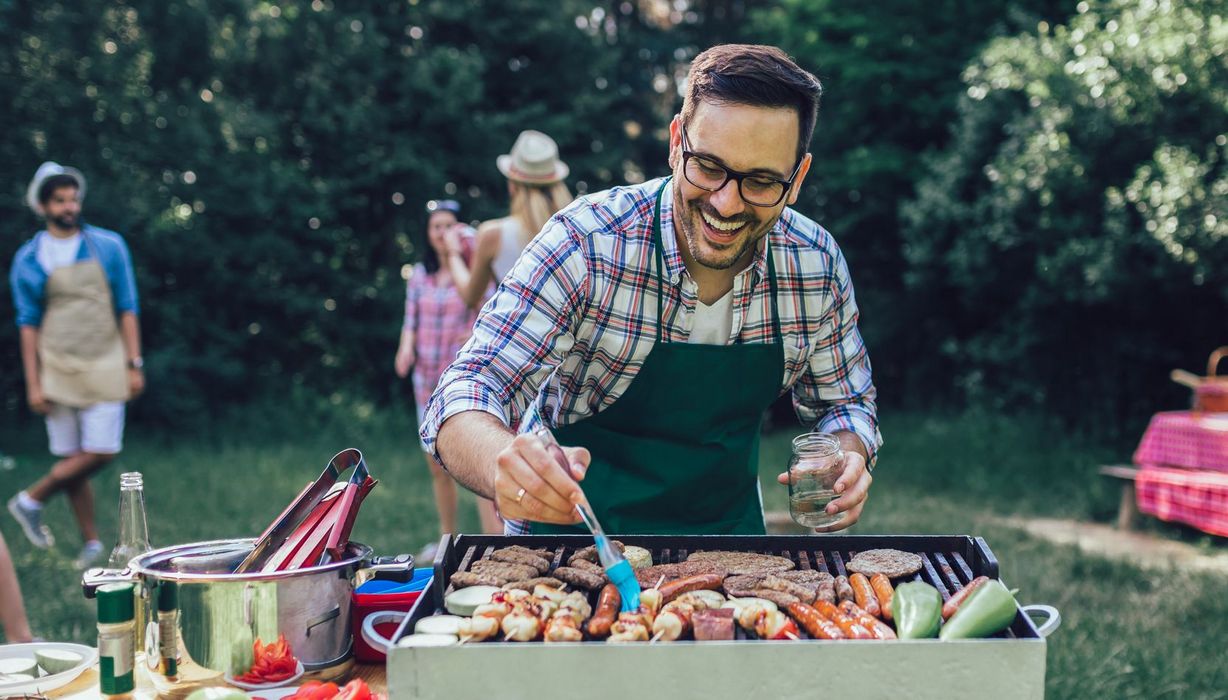 Mann bestreicht freudestrahlend einen Gemüsespies, der gemeinsam mit Zuchinischeiben, Paprika, Würstchen und Burgerpatties auf einem Grill liegt. Im Hintergrund sind unscharf ein paar gutgelaunte Personen zu sehen.