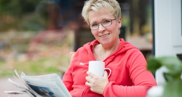 Frau im mittleren Alter sitzt mit einer Tasse Kaffee und einer Zeitung auf einer Terrasse und blickt in die Kamera