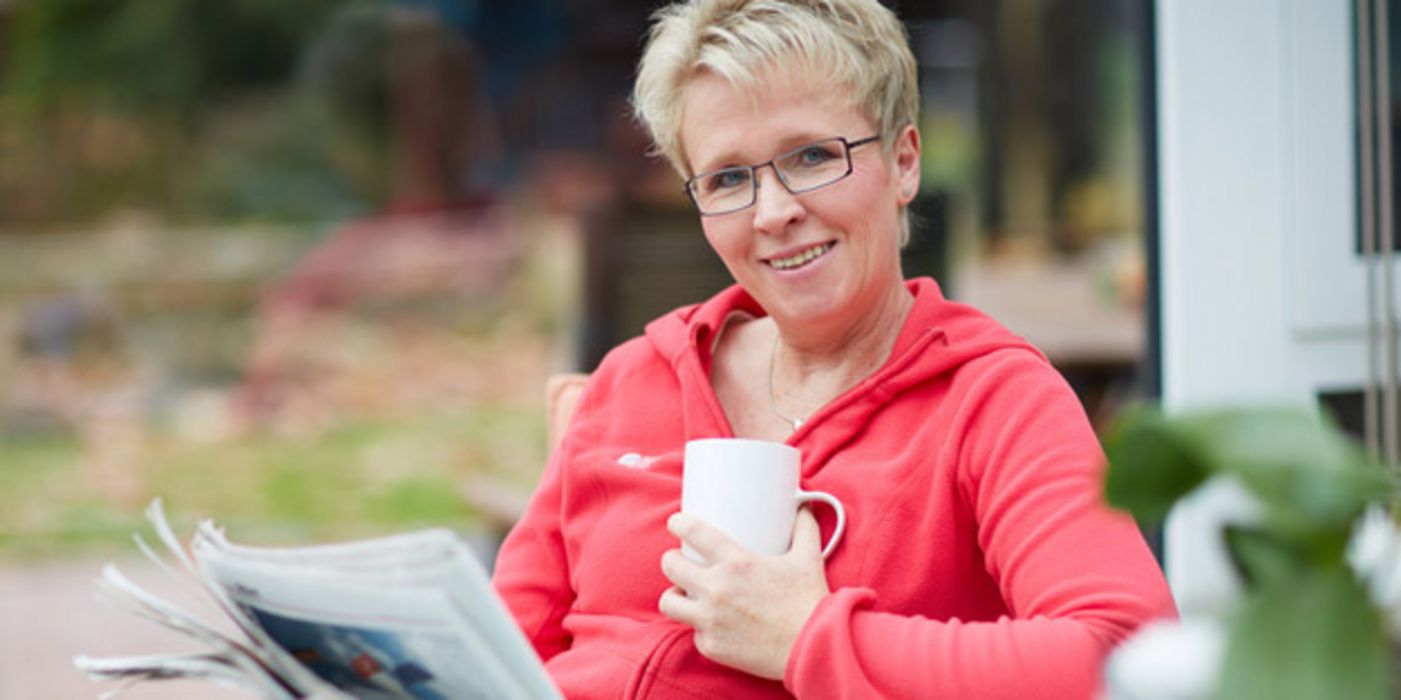 Frau im mittleren Alter sitzt mit einer Tasse Kaffee und einer Zeitung auf einer Terrasse und blickt in die Kamera