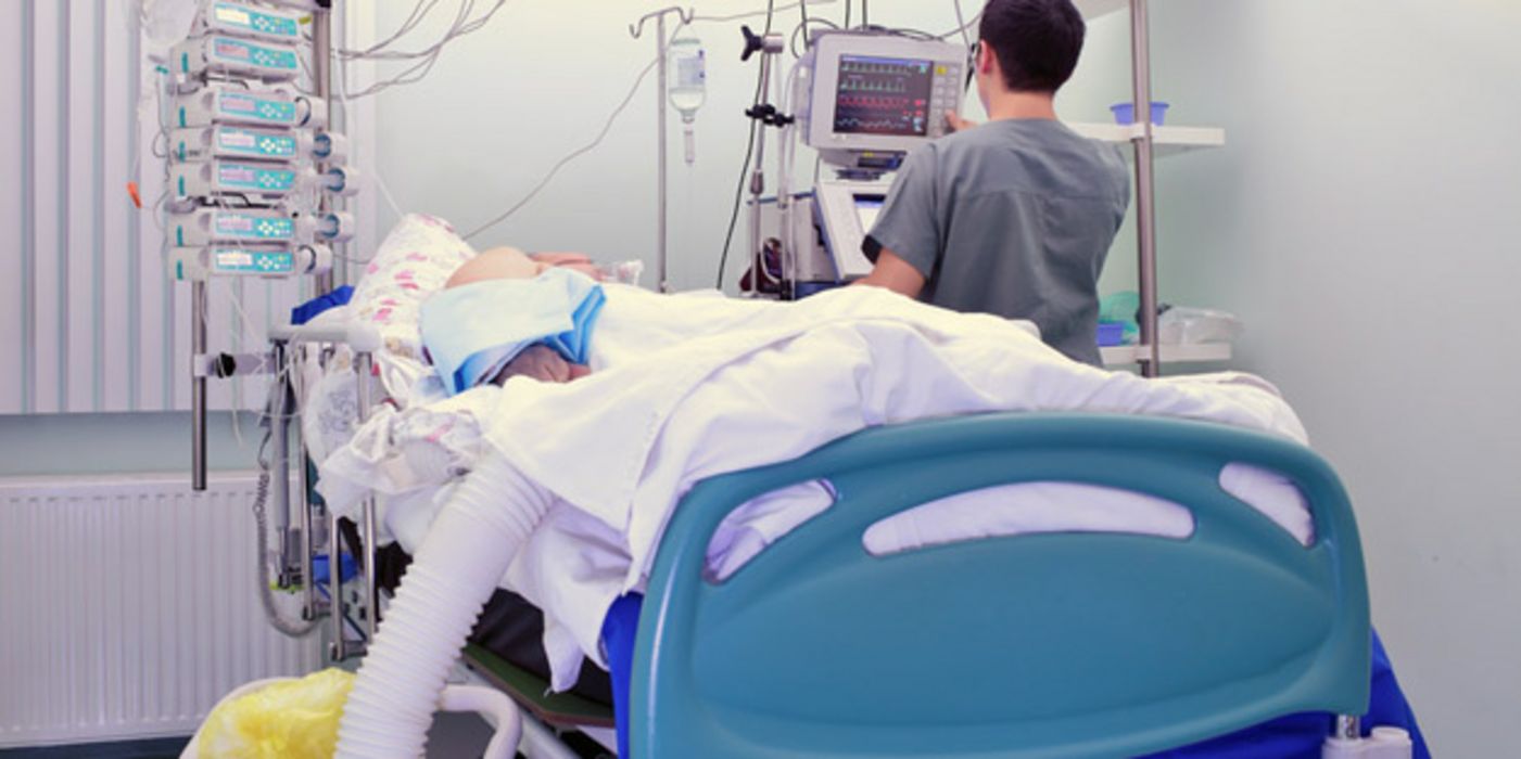 Intensivstation, Bett mit Patient, vom Fußende aus fotografiert, unkenntlich, Pfleger am Kopfenede neben Bett (Rückansicht) an Apparaturen
