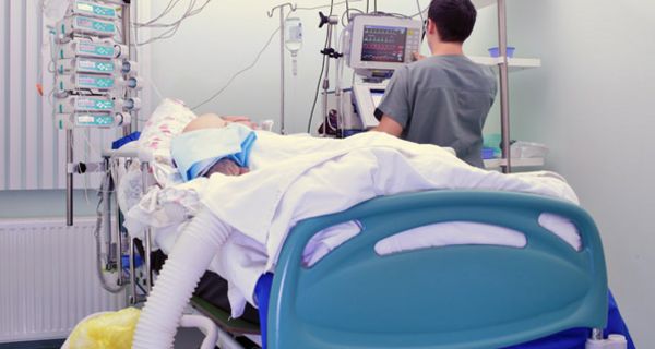 Intensivstation, Bett mit Patient, vom Fußende aus fotografiert, unkenntlich, Pfleger am Kopfenede neben Bett (Rückansicht) an Apparaturen
