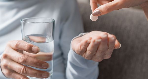 Ältere Dame öffnet eine Hand, um eine Tablette entgegen zu nehmen. In der anderen Hand hält sie ein Glas Wasser. 