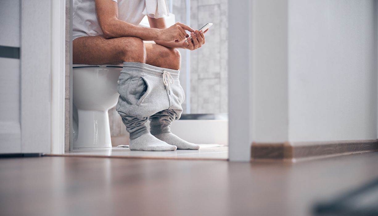 Mann, sitzt mit Handy auf der Toilette.