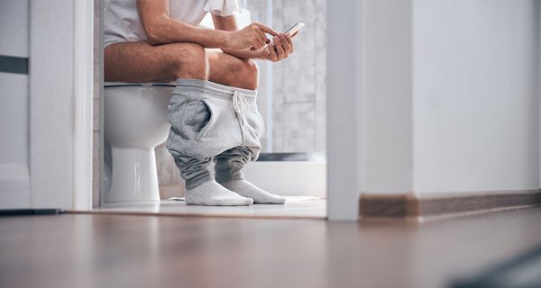 Mann, sitzt mit Handy auf der Toilette.
