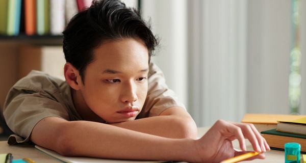 Junge am Schreibtisch liegt mit den Armen auf einem Heft. Er spielt mit einem Bleistift und einem Spitzer.