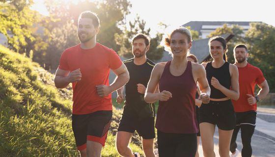 Zwei gutelaunte Frauen und drei Männer joggen bei schönem Wetter auf einem asphaltierten Weg.