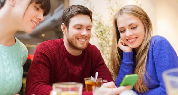 Junge Leute um die 20 (2 Frauen, 1 Mann), am Tisch sitzend, schauen lachend in grünes Smartphone, das der Mann in der Hand hält