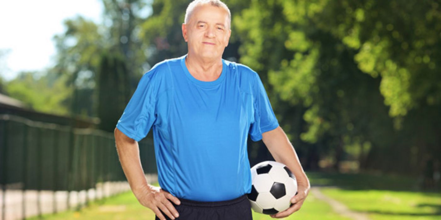 Mann in den 60ern, grauhaarig, blaues Fußballtrikot, schwarze Hose, Ball unterm Arm, eine Hand in der Hüfte, Hintergrund Park