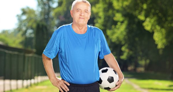 Mann in den 60ern, grauhaarig, blaues Fußballtrikot, schwarze Hose, Ball unterm Arm, eine Hand in der Hüfte, Hintergrund Park