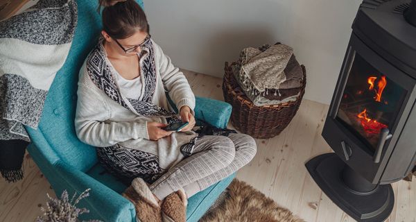 Junge Frau, sitzt im Winter auf einem Sessel vor dem Kamin, mit Handy in der Hand.