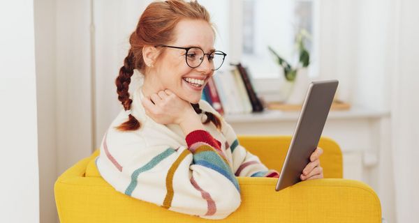 Junge Frau, sitzt auf der Couch und schaut lächelnd auf ihr Tablet.