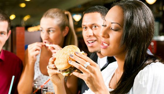 2 Paare, 1 Afro-amerikanisches, im Vordergrund, Frau beißt genüsslich in Hamburger, andere essen ebenfalls irgend welches Fastfood
