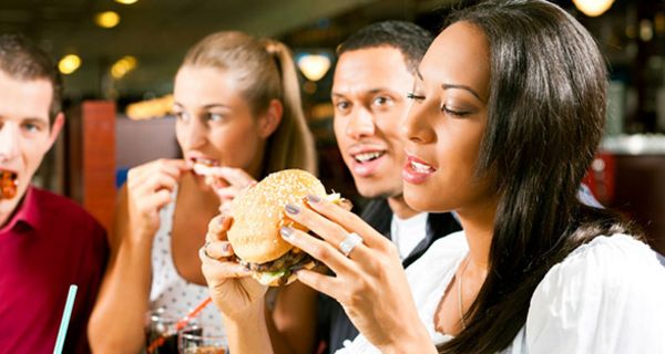 2 Paare, 1 Afro-amerikanisches, im Vordergrund, Frau beißt genüsslich in Hamburger, andere essen ebenfalls irgend welches Fastfood