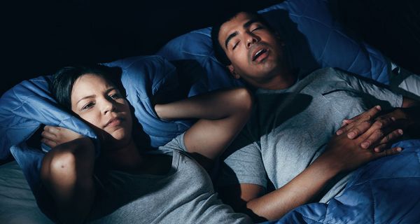 Junges Paar im Bett, Mann schnarcht, Frau hält sich die Ohren zu.