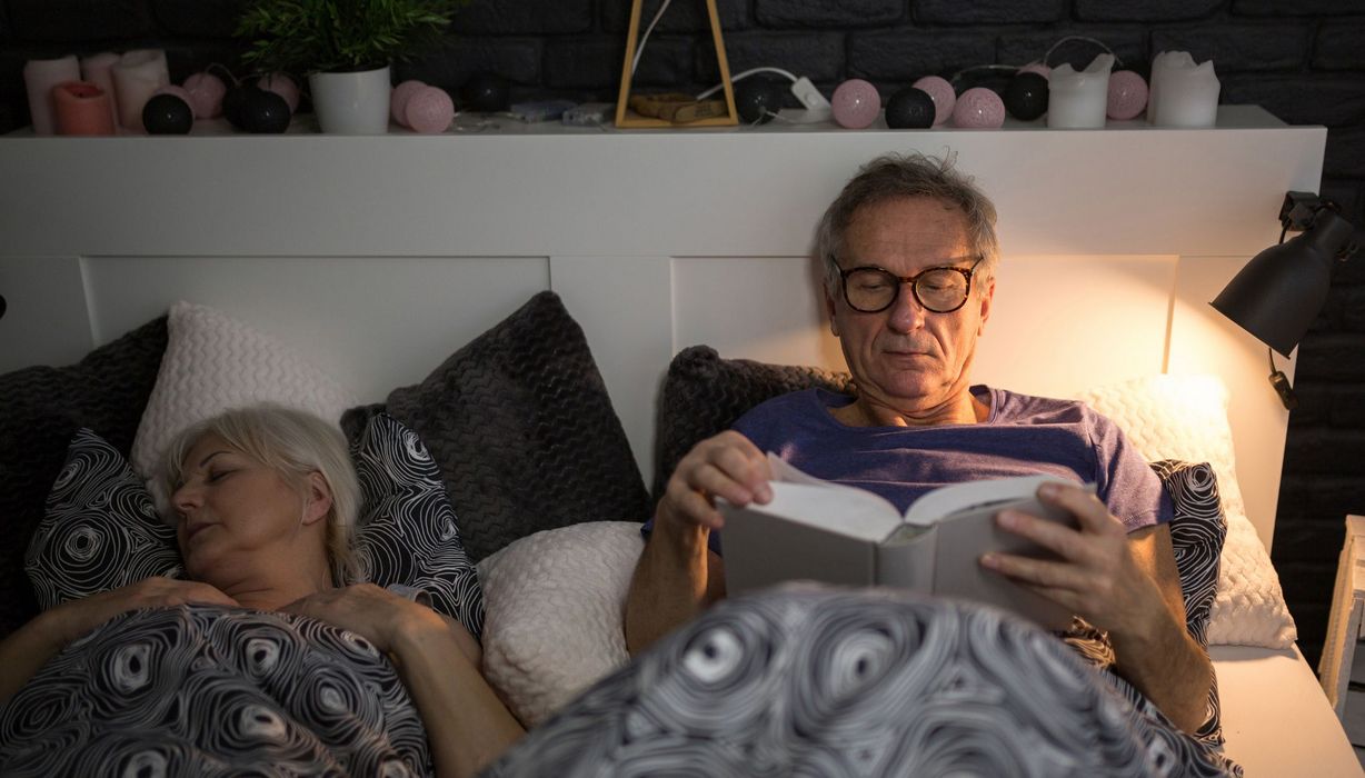 Senioren-Paar liegt im Bett, Mann ist noch wach und liest.