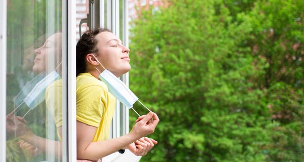 Junge Frau, lehnt sich aus dem Fenster und zieht Mund-Nasen-Schutz ab.