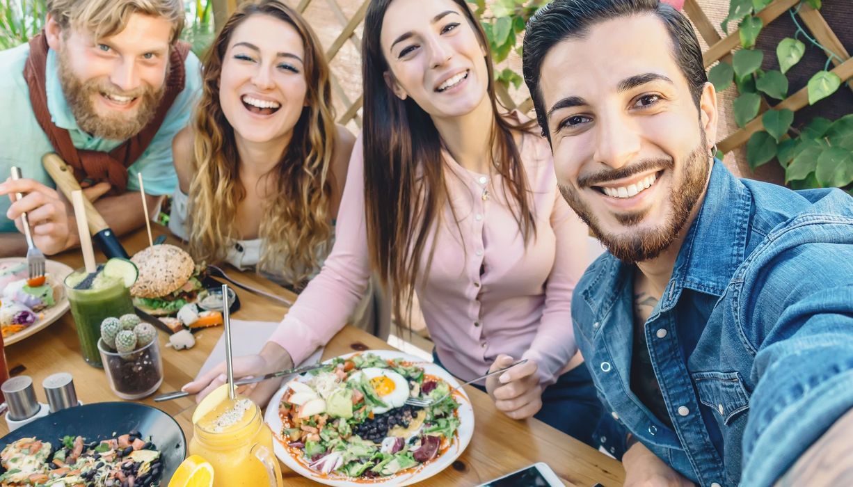 Vier junge Menschen sitzen um einen Tisch mit vegetarischem essen und machen ein Selfie.