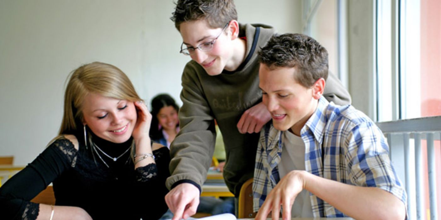 Schülerin und zwei Schüler im Klassenzimmer, ca. 15 Jahre alt, schauen gemeinsam in Unterlagen