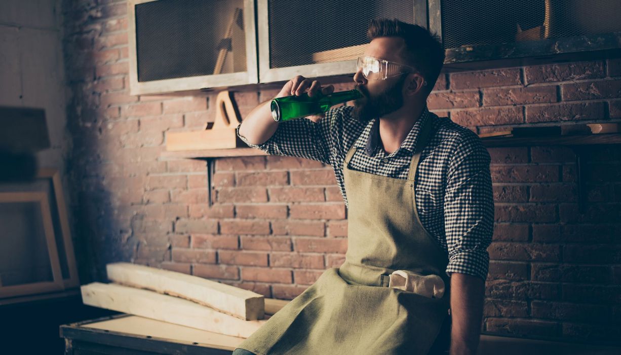 Handwerker, sitzt auf Arbeitsbank und trinkt ein Bier.