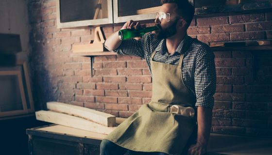 Handwerker, sitzt auf Arbeitsbank und trinkt ein Bier.