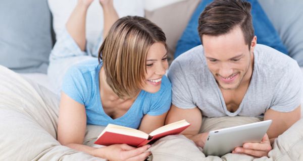 Junges Paar liegen auf der Couch auf dem Bauch gemütlich beim Lesen: Sie mit Buch, er mit E-Book-Reader