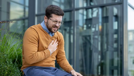 Mann zwischen 30 und 40 Jahren sitzt vor einem Bürogebäude auf einer Bank und hält sich die Brust.