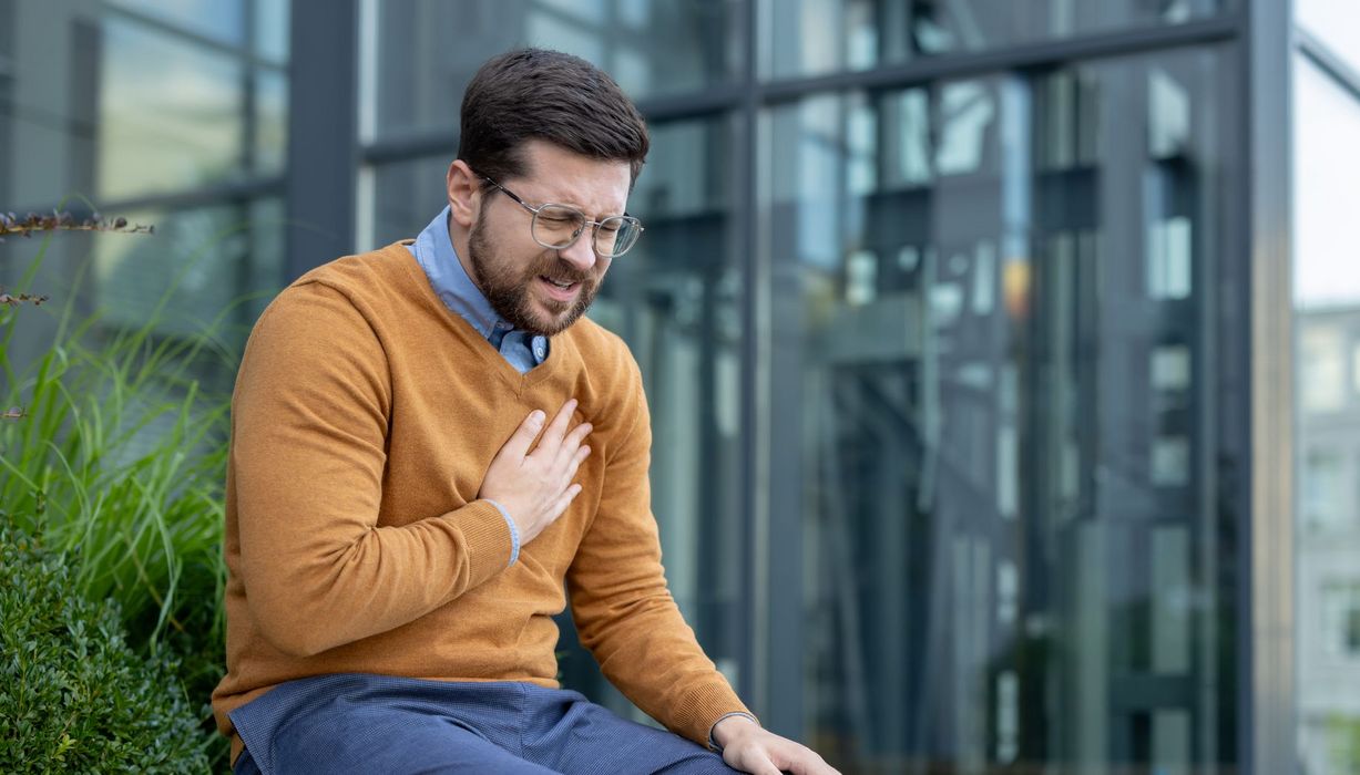 Mann zwischen 30 und 40 Jahren sitzt vor einem Bürogebäude auf einer Bank und hält sich die Brust.