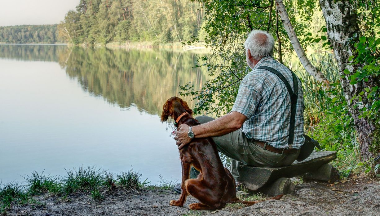 Älterer Mann, sitzt mit Hund am See.