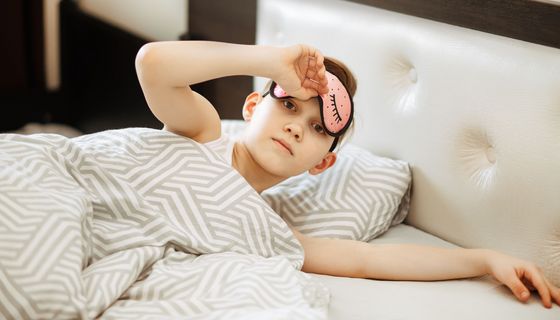Ein Mädchen liegt im Bett und lugt unter einer Schlafmaske hervor.