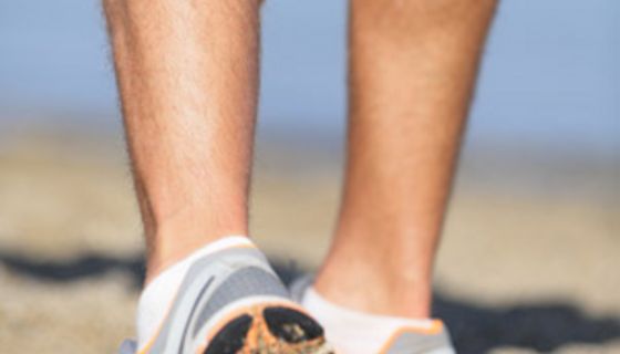 Mensch in Turnschuhen am Strand greift sich an die linke Wade.