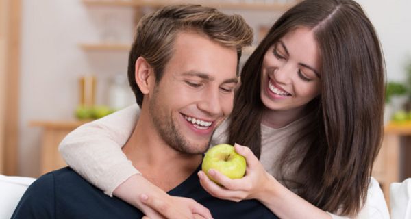 Frau und Mann um die 30, Frau hält lachend dem ebenfalls lachenden Mann einen Apfel zum Anbeißen hin