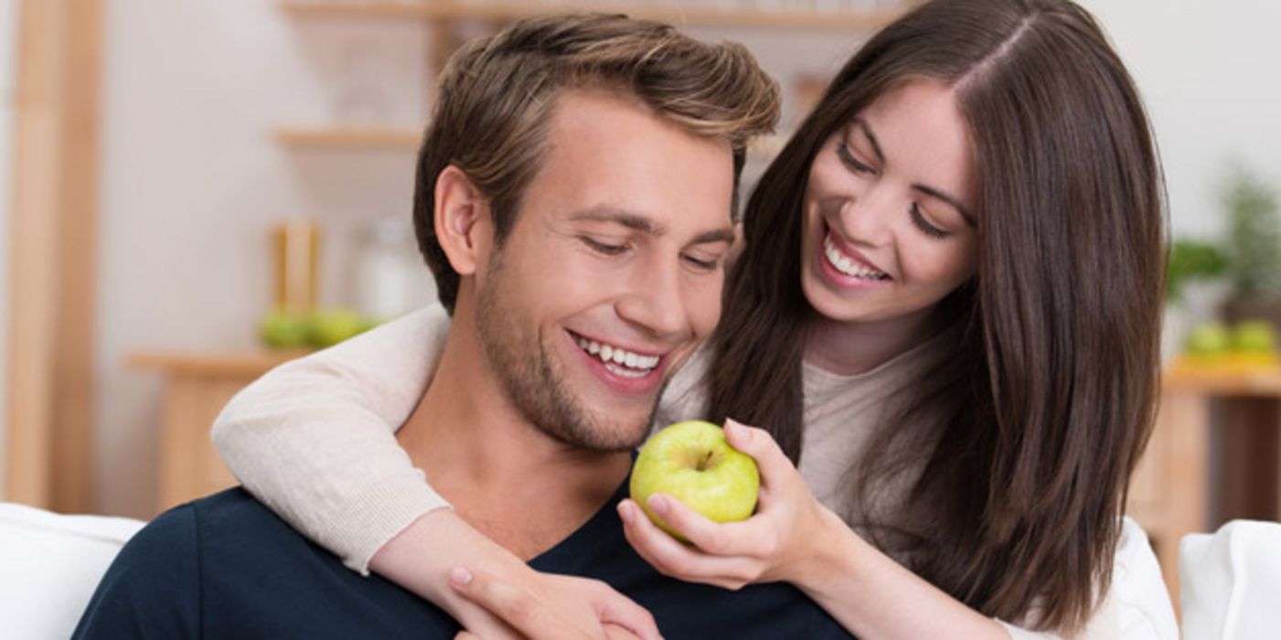 Frau und Mann um die 30, Frau hält lachend dem ebenfalls lachenden Mann einen Apfel zum Anbeißen hin