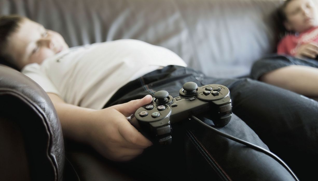 Übergewichtiger Junge mit Gaming-Controller auf der Couch.