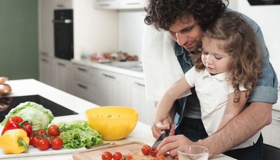Mann hat ein kleines Mädchen, etwa drei Jahre alt, im Arm. Sie schneiden gemeinsam Tomaten für einen Salat.