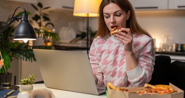Frau sitzt abends vor dem Laptop und ist ein Stück Pizza.